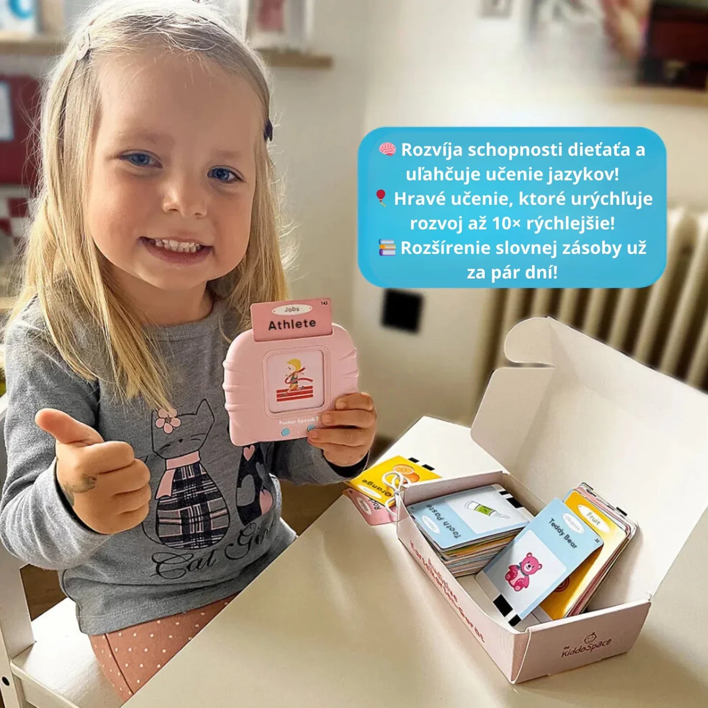 O fetiță fericită oferind un semn de apreciere în timp ce folosește dispozitivul roz Kinderspeech, având în față cutia cu carduri educaționale pentru dezvoltarea rapidă a limbajului și a vocabularului prin joc interactiv