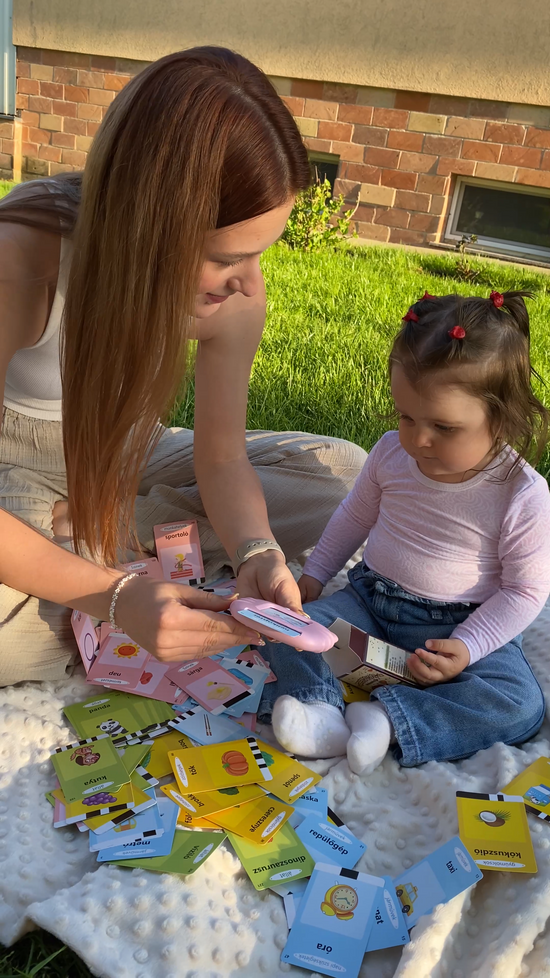 O mamă și fetița ei așezate pe o pătură în aer liber, folosind dispozitivul educațional roz Kinderspeech pentru a explora o multitudine de carduri colorate, o activitate interactivă menită să sprijine dezvoltarea limbajului într-un mediu relaxat și natural