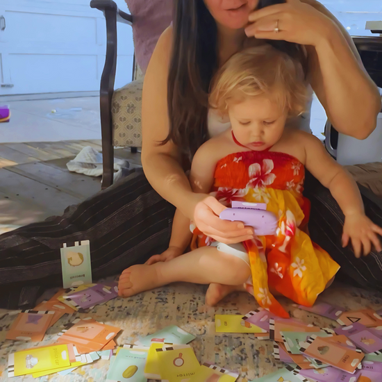 O mamă și fetița ei așezate pe podea, explorând împreună o multitudine de carduri educaționale și folosind dispozitivul Kinderspeech pentru a învăța cuvinte noi și a stimula dezvoltarea timpurie a limbajului prin interacțiune și joc