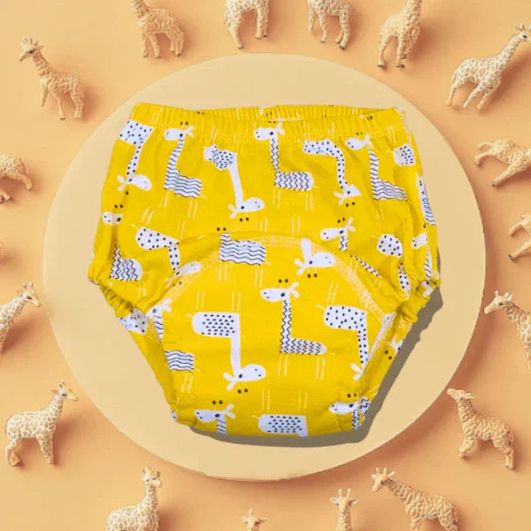 O pereche de chiloței de antrenament pentru copii, de culoare galben intens cu imprimeu vesel cu girafe, prezentați pe un fundal decorativ înconjurați de figurine mici sub formă de girafă, ideali pentru tranziția distractivă de la scutece