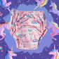 O pereche de chiloței de antrenament pentru copii, de culoare roz cu un imprimeu magic cu unicorni și curcubeie, prezentați pe un fundal mov decorat cu ilustrații de unicorni, ideali pentru a face tranziția la oliță o experiență de poveste