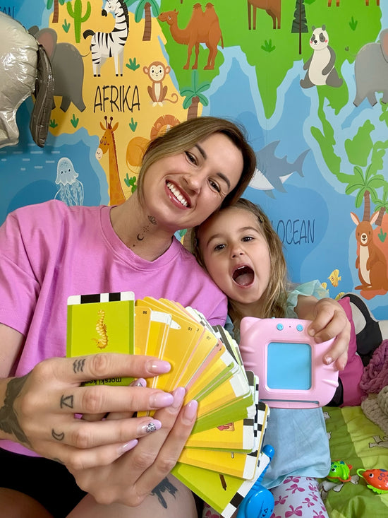 O mamă și fetița ei zâmbind fericite în timp ce prezintă un set bogat de carduri educaționale colorate și dispozitivul roz Kinderspeech, ilustrând o experiență de învățare interactivă și distractivă într-un decor vesel cu hărți și animale